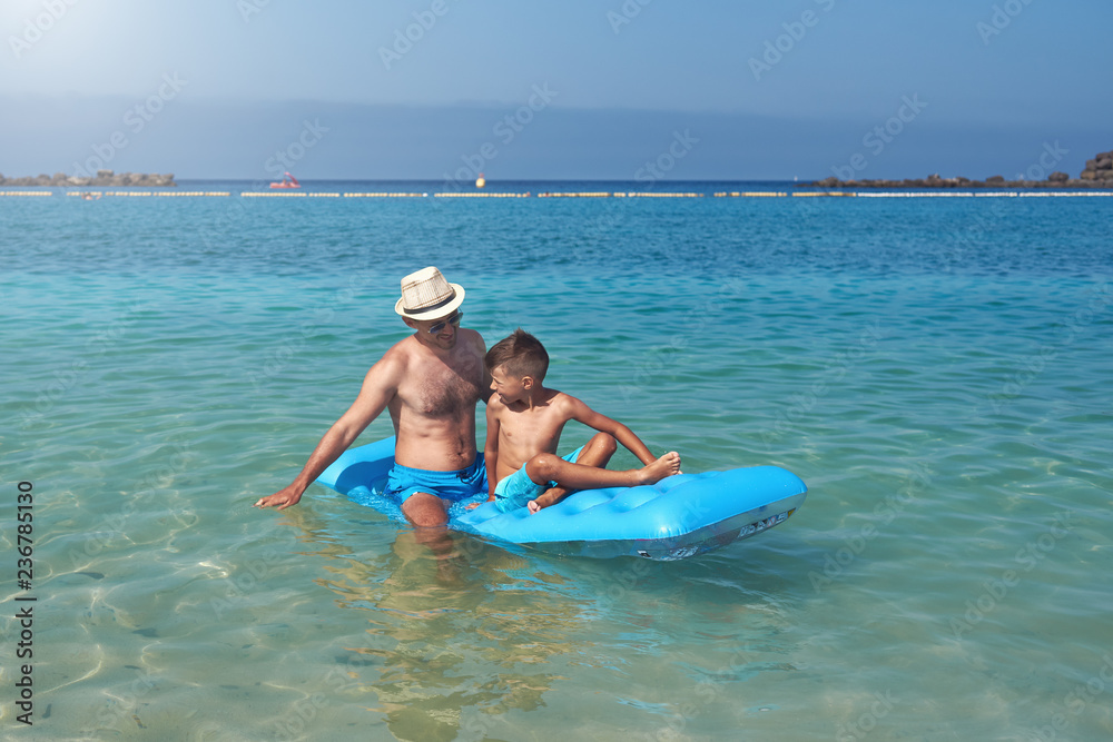 Dad and son are sitting on a blue inflatable floater in the clear ocean water. They are looking to each other, having fun and enjoying their vacations.