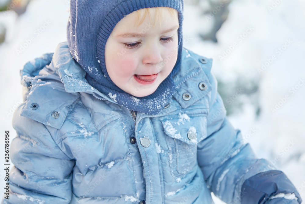 Cute caucasian liittle boy with bright blue eyes in winter clothes and hat (hood) on winter, with tongue outside. Healthy childhood. Outdoors, winter activity. Copy space, close up portrait.