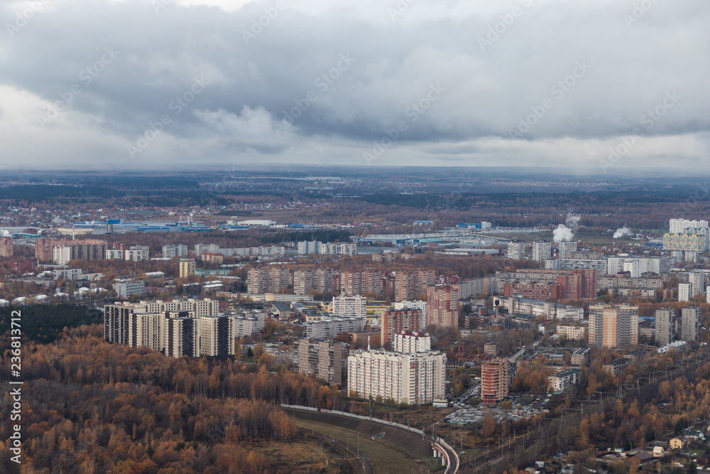 High-rise buildings near the road. A small city with an altitude of the aircraft. At the top of the white clouds on the horizon, below the forest with the road, city buildings.