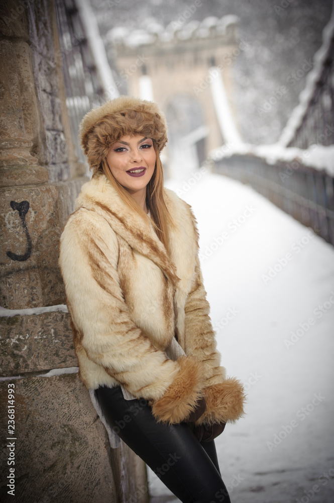 Attractive woman with brown fur cap and jacket enjoying the winter. Side view of fashionable blonde girl posing against snow covered bridge. Beautiful young female with cold weather outfit Attractive woman with brown fur cap and jacket enjoying the winter. Side view of fashionable blonde girl posing against snow covered bridge. Beautiful young female with cold weather outfit