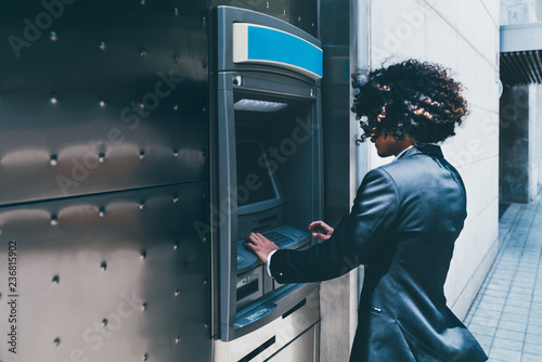 Side view of a businessman withdrawing money from his bank card using an outdoor ATM;  a curly man entrepreneur is using automated teller machine on the street to to replenish his account deposit