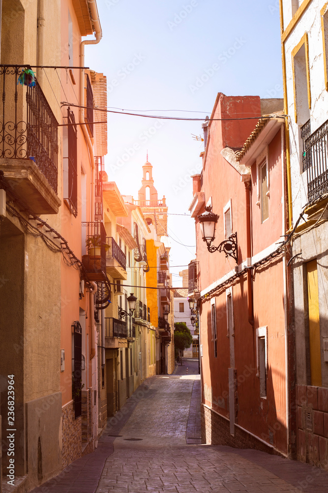 Fototapeta premium The church can be seen at the end of the cozy street of the old town of Releu in the rays of the sun. Mediterranean architecture in Spain.