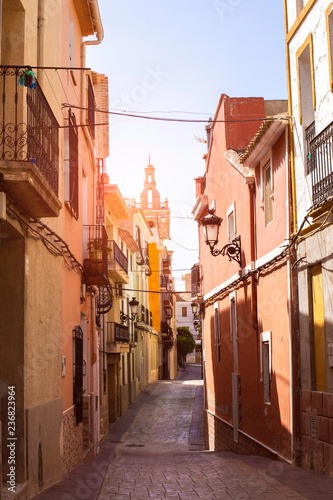 The church can be seen at the end of the cozy street of the old town of Releu in the rays of the sun. Mediterranean architecture in Spain.