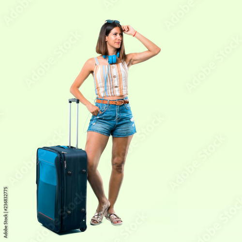 Full body of Girl traveling with her suitcase having doubts and with confuse face expression while scratching head on isolated green background