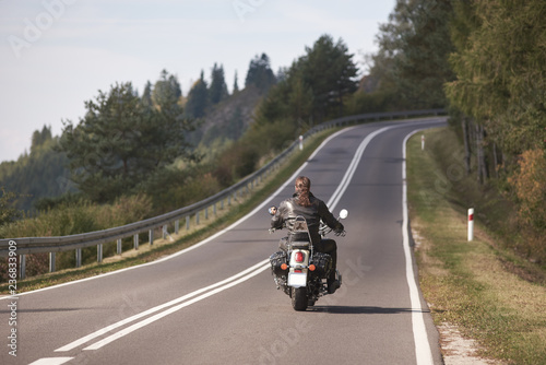 Back view of biker in black leather outfit with long hair riding motorbike up twisted empty asphalt road on bright sunny summer day by green forest trees under clear cloudless sky.
