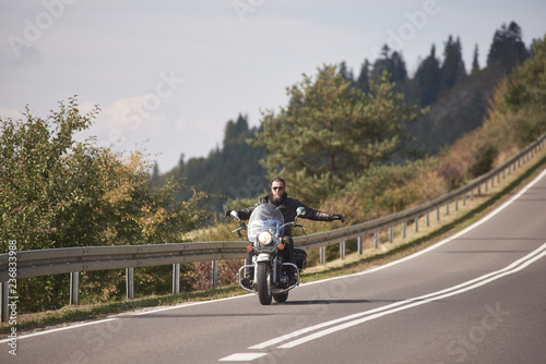 Alone motorcyclist in black leather outfit riding cruiser bike with stretched out arms on bright sunny summer day on background of foggy distant green woody hills under morning sky.