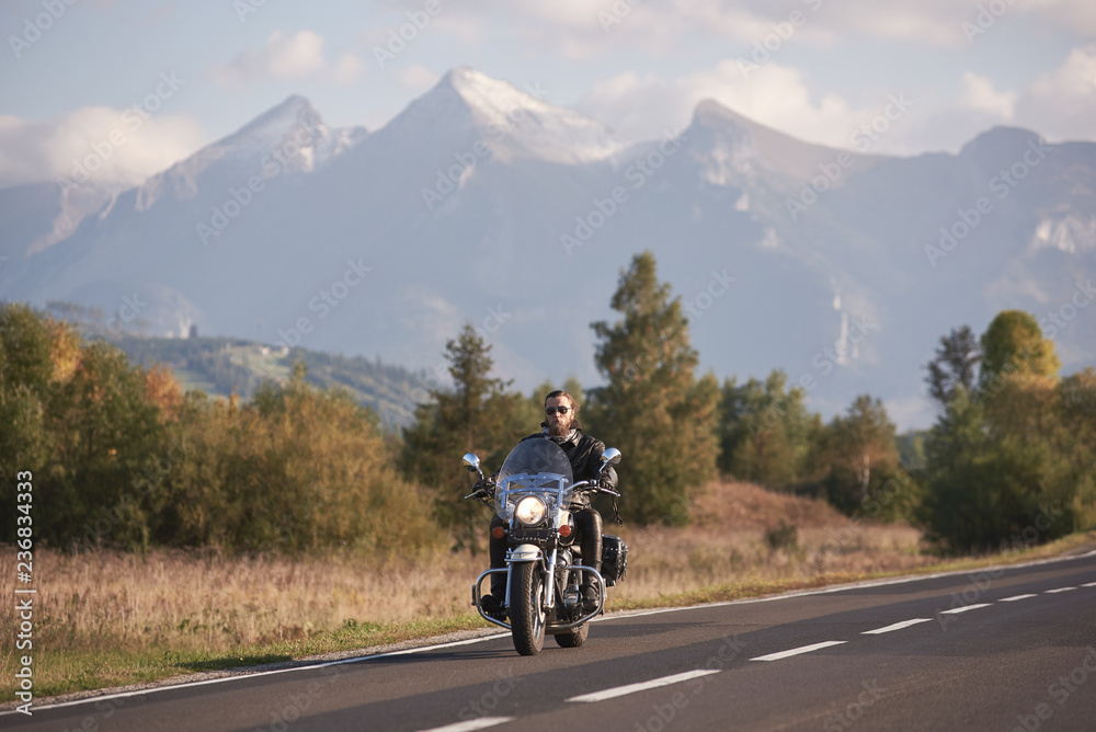 Bearded biker in sunglasses and black leather clothing riding modern powerful cruiser motorbike on blue foggy mountains with white snowy peaks and bright autumn sky copy space background.