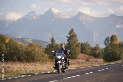 Bearded biker in sunglasses and black leather clothing riding modern powerful cruiser motorbike on blue foggy mountains with white snowy peaks and bright autumn sky copy space background. © anatoliy_gleb