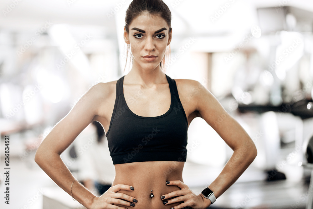 Beautiful slim girl dressed in black sportswear is standing in the gym and holding her hands on her waist