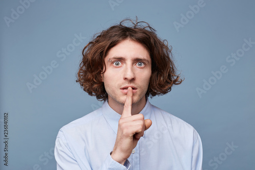 Serious frightened male with long messy hair, keeps fore finger on lips, tries to keep conspiracy, looks camera, says: Shh, make silence please. Isolated shot of man shows silence gesture photo