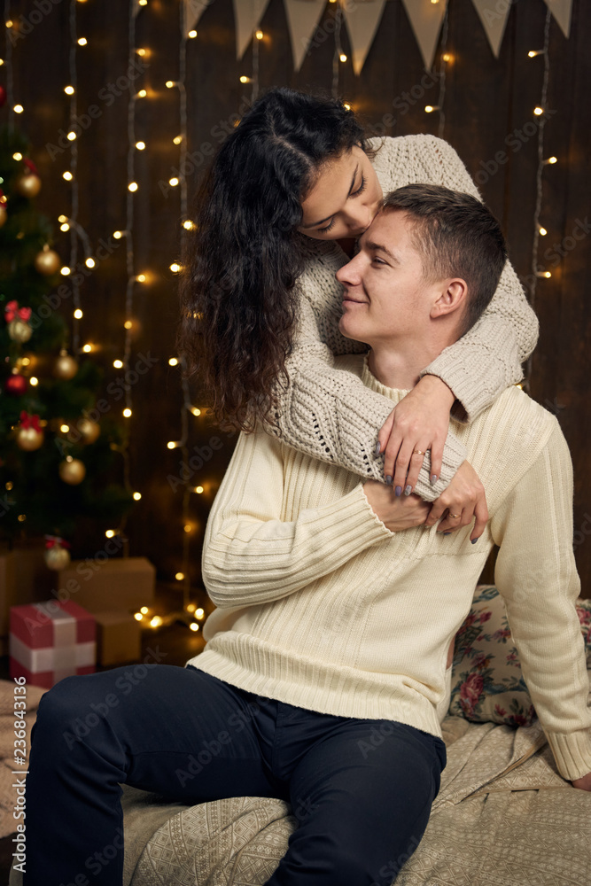young couple is in christmas lights and decoration, dressed in white, fir tree on dark wooden background, winter holiday concept young couple is in christmas lights and decoration, dressed in white, fir tree on dark wooden background, winter holiday concept