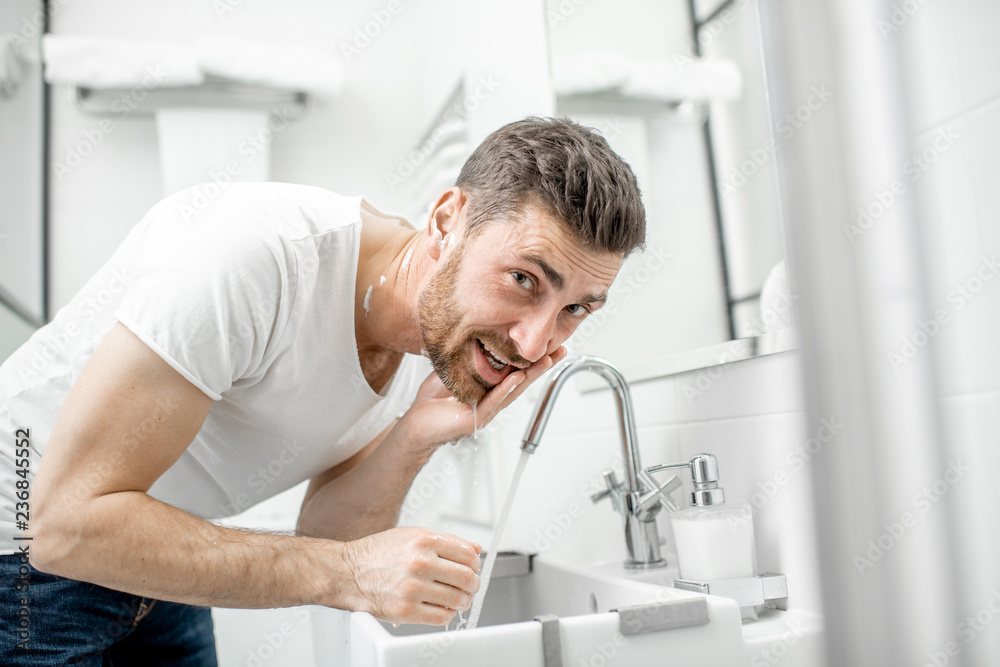 Man washing his face with fresh water and foam in the sink at the white bathroom