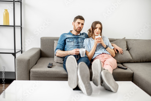 Young couple with unhappy faces watching boring TV sitting together on the couch at home