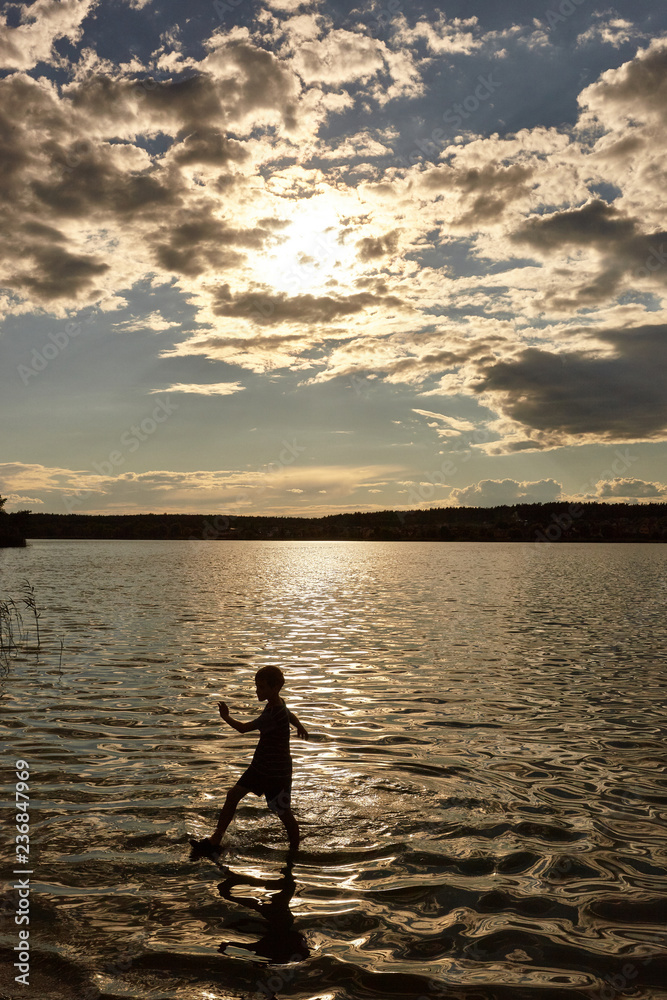 Silhouette of a boy walking on the water in the rays of the sunset. Beach by the sea, lake or river