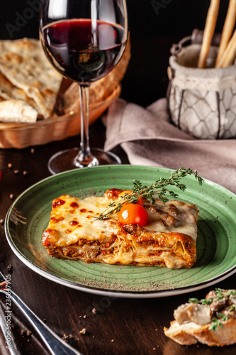 The concept of Italian cuisine. Baked lasagna with minced bolognese, pasta, cherry tomatoes lies on a green plate in a restaurant. A glass of red wine and bread basket on the table