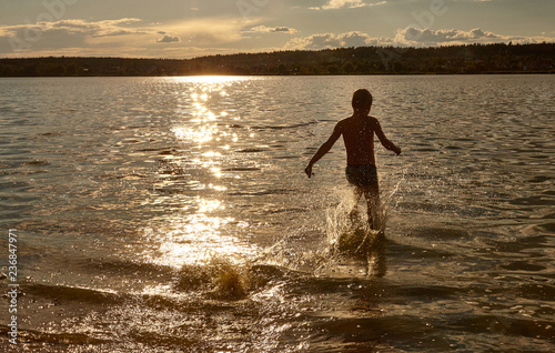Silhouette of a boy running through the water in the rays of the sunset. Beach and splashing water by the sea, lake or river