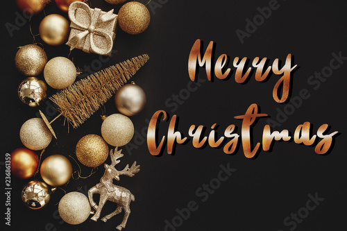 Merry Christmas text, handwritten golden sign at christmas golden gift box, tree, reindeer and glitter baubles on stylish black background. Seasons greetings card