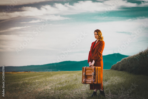 Portrait of young woman in red coar at autumn countryside with suitcase. photo
