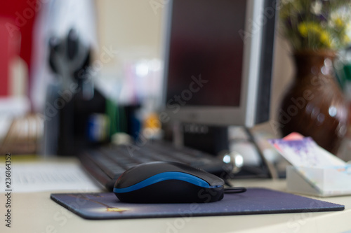 Small digital mouse on the table. On background is desktop monitor, bouquet of flowers and keyboard