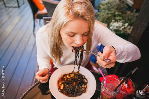 Young beautiful woman eating black pasta with seafood and cuttlefish ink in the outdoor restaurant. Funny and beautiful.