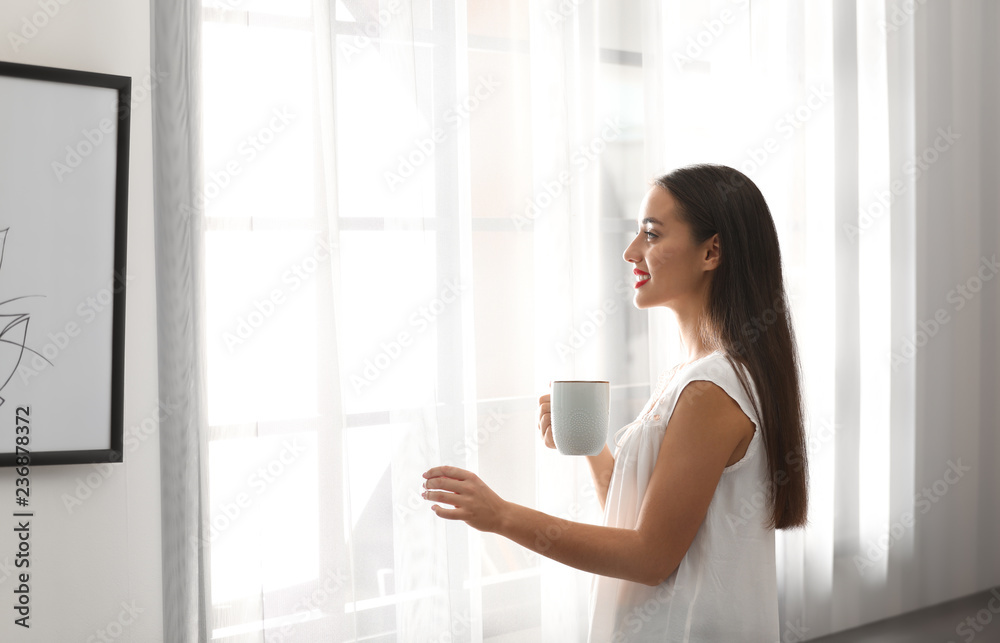 Young woman near window with curtains at home. Space for text Young woman near window with curtains at home. Space for text