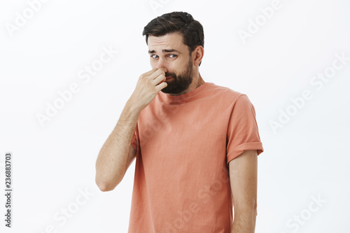 Gosh it stinks. Disgusted and displeased unhappy male with beard closing nose with fingers looking at creepy and awful object which reeks with terrible smell looking at lower right corner dissatisfied photo