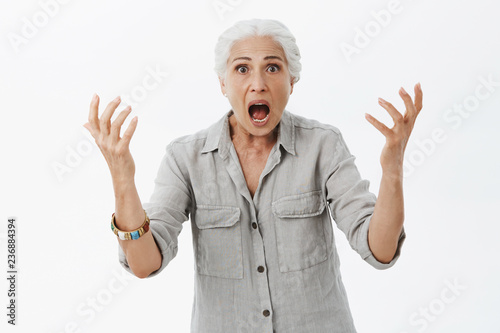 Waist-up shot of excited and shocked confused pissed elderly mother shaking with raised arms opening mouth from surprise and worry arguing seeing stupid and shocking action of son over gray wall