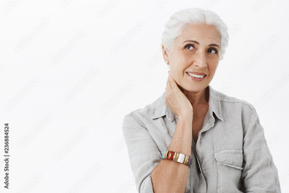 Waist-up shot of gentle delighted european senior female with white hair touching neck and smiling joyfully gazing at upper left corner feeling healthy and energized even in old age Waist-up shot of gentle delighted european senior female with white hair touching neck and smiling joyfully gazing at upper left corner feeling healthy and energized even in old age