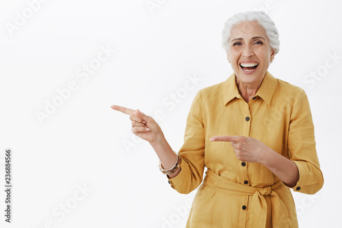 Portrait of delighted healthy and active pretty senior female in yellow trendy coat and bracelet laughing out loud from amusement standing entertained and charistmatic pointing left with forefingers photo