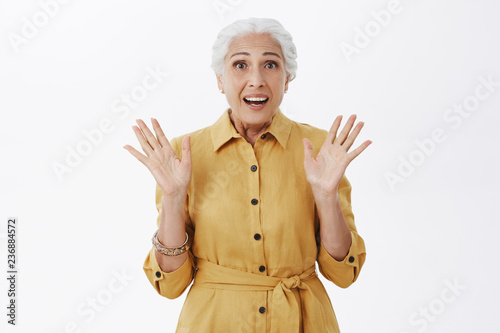 Portrait of excited and surpised fashionable grandmother with white hair in yellow trendy coat raising palms in amazement gazing at awesome sale feeling astonished over gray background photo