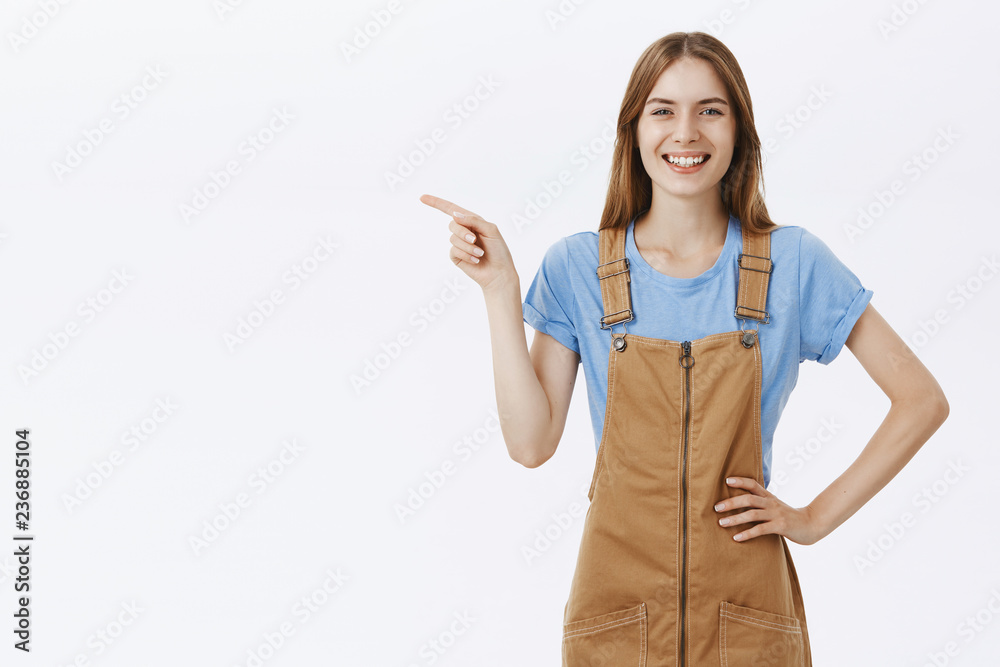 Portrait of friendly-looking pleased and carefree charming woman in brown overalls over blue t-shirt holding hand on hip pointing left and smiling joyfully showing perfect copy space against grey wall Portrait of friendly-looking pleased and carefree charming woman in brown overalls over blue t-shirt holding hand on hip pointing left and smiling joyfully showing perfect copy space against grey wall