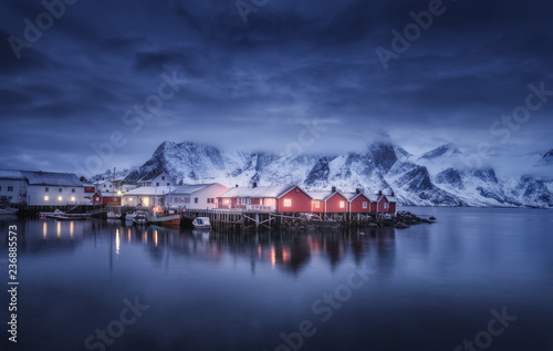 Beautiful fishing village with boats at night, Lofoten islands, Norway. Winter landscape with houses, illumination, snowy mountains, sea, blue cloudy sky reflected in water at dusk. Norwegian rorbuer
