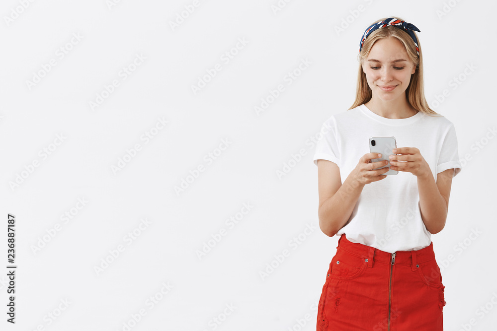 Girl melting from sweet messages received from boyfriend. Portrait of cute happy young caucasian blonde in red skirt and headband, holding smartphone, smiling and gazing at device screen tender Girl melting from sweet messages received from boyfriend. Portrait of cute happy young caucasian blonde in red skirt and headband, holding smartphone, smiling and gazing at device screen tender