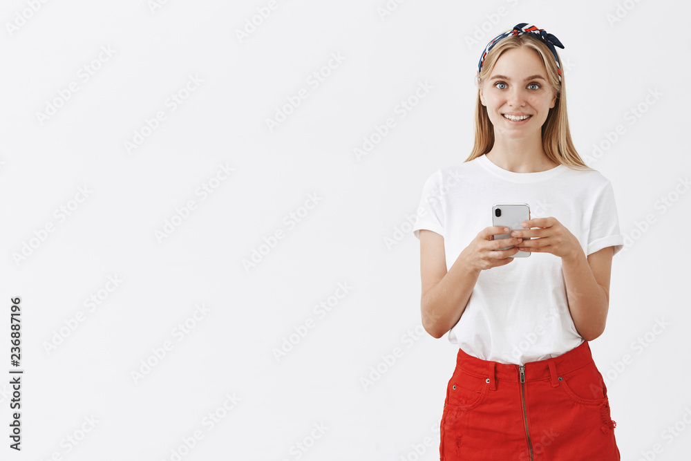 Indoor shot of happy good-looking blonde female in red skirt and headband looking happily at camera holding smartphone making notes during interview with gadget over gray background Indoor shot of happy good-looking blonde female in red skirt and headband looking happily at camera holding smartphone making notes during interview with gadget over gray background