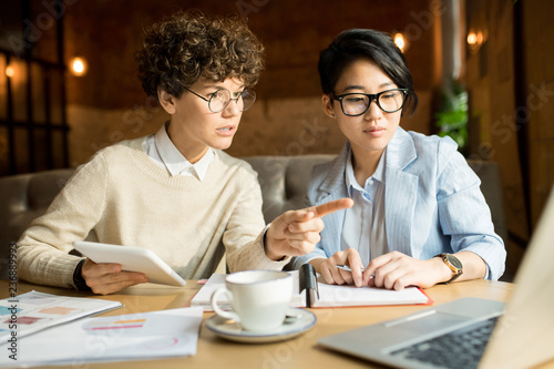 Serious pensive young marketers in glasses sitting at table and discussing strategy: curly-haired lady offering idea and pointing at laptop screen photo