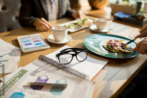 Close-up of unrecognizable women sitting at table with glasses, notepad and marketing charts and eating tasty dinner while taking break during business lunch