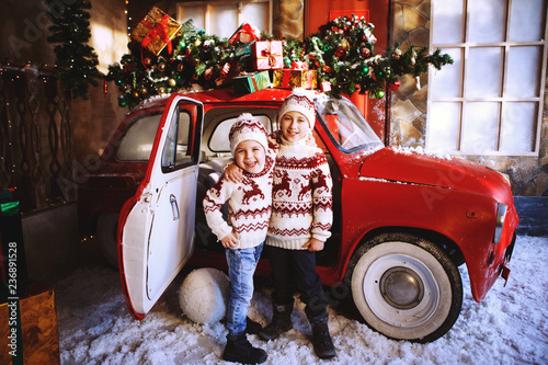Two brothers in knitted sweaters with deer stand near the red Christmas car and rejoice at the advent of Christmas.