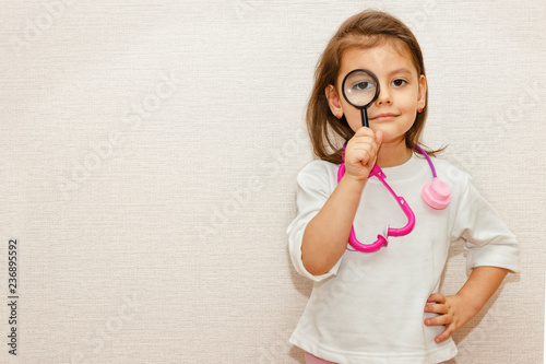 Cute little girl is playing doctor with magnifier and stethoscope on beige background.