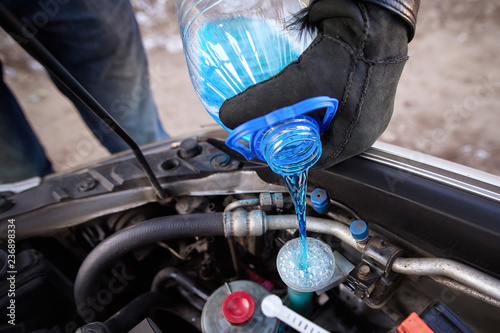 Hands in gloves holding bottle with non-freezing washer fluid and pouring it into windshield washer tank in winter. Replacement technical fluids according to season. Washer reservoir completely filled photo