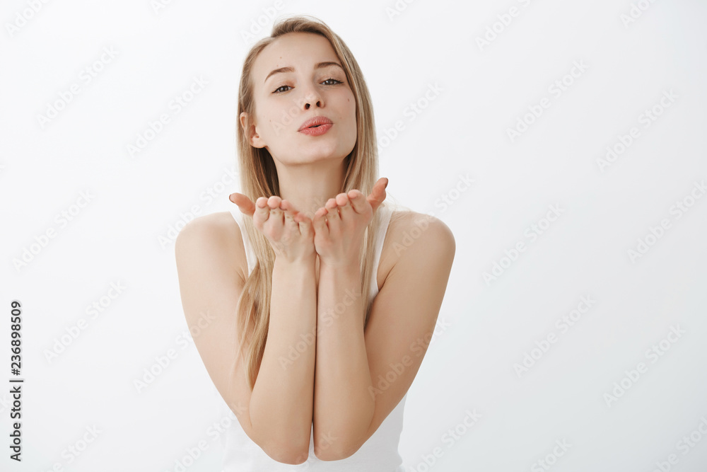 Tender and flirty young attractive female with blond hair in tank-top raising hands near folded lips as blowing kiss at camera with sympathy and love in eyes, smiling sensually over gray wall Tender and flirty young attractive female with blond hair in tank-top raising hands near folded lips as blowing kiss at camera with sympathy and love in eyes, smiling sensually over gray wall