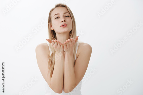 Tender and flirty young attractive female with blond hair in tank-top raising hands near folded lips as blowing kiss at camera with sympathy and love in eyes, smiling sensually over gray wall