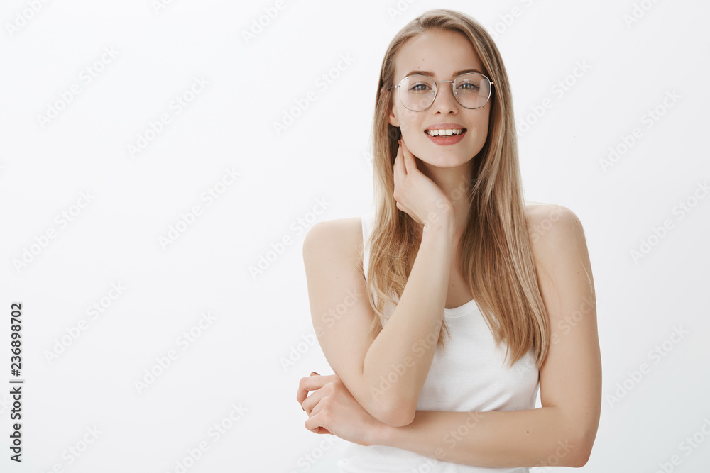 Indoor shot of sensual charming slender female blonde in transparent glasses touching neck shy and gentle as smiling friendly at camera, flirting making gazes at camera as posing over gray wall Indoor shot of sensual charming slender female blonde in transparent glasses touching neck shy and gentle as smiling friendly at camera, flirting making gazes at camera as posing over gray wall