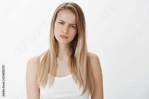 Girl suspects something wrong being doubtful in friend telling whole truth, squinting suspicious at camera as frowning and making hesitant face, standing questioned and intense over gray wall