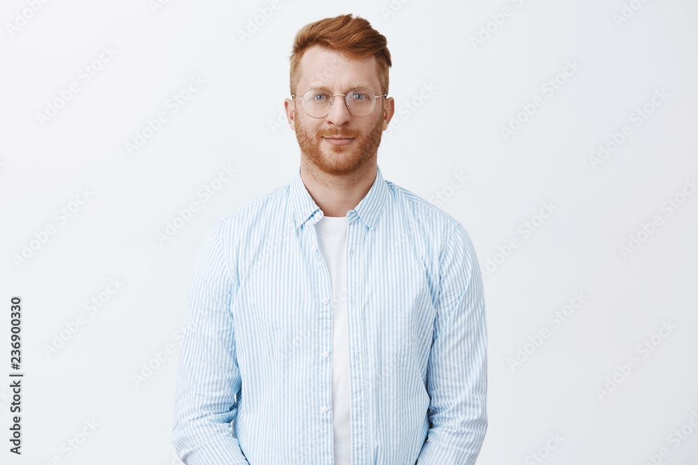 Good-looking creative and smart redhead male entrepreneur hiring new employees, standing in casual shirt and glasses over gray wall, smiling friendly, gazing at camera with thoughtful, determined look Good-looking creative and smart redhead male entrepreneur hiring new employees, standing in casual shirt and glasses over gray wall, smiling friendly, gazing at camera with thoughtful, determined look