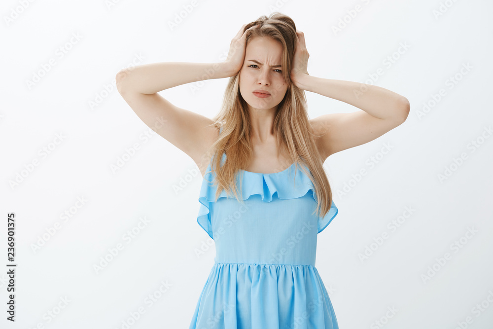 Moody displeased good-looking young female with fair hair in blue dress frowning grabbing head and squinting at camera suspicious and doubtful, having hangover after cool party over grey wall Moody displeased good-looking young female with fair hair in blue dress frowning grabbing head and squinting at camera suspicious and doubtful, having hangover after cool party over grey wall