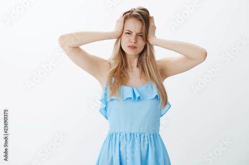 Moody displeased good-looking young female with fair hair in blue dress frowning grabbing head and squinting at camera suspicious and doubtful, having hangover after cool party over grey wall © Cookie Studio