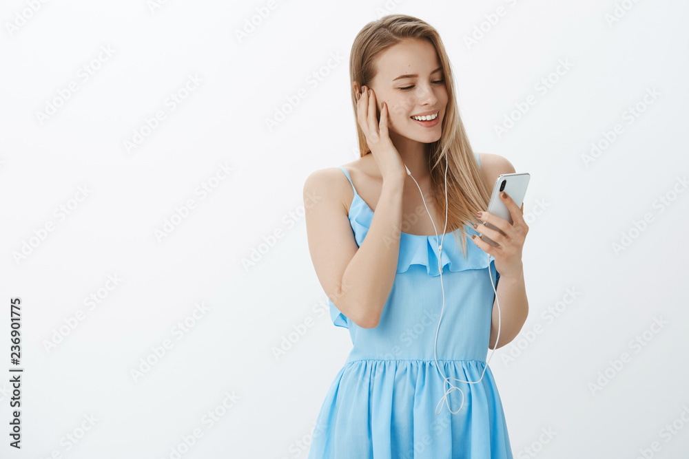 Studio shot of young attractive and glamour blonde female in blue dress enjoying listening music in earphones touching earbud, smiling broadly at cellphone screen, liking new album of favorite singer Studio shot of young attractive and glamour blonde female in blue dress enjoying listening music in earphones touching earbud, smiling broadly at cellphone screen, liking new album of favorite singer