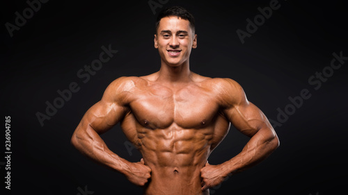 Strong smiley fitness model showing muscles over dark background. © Stavros