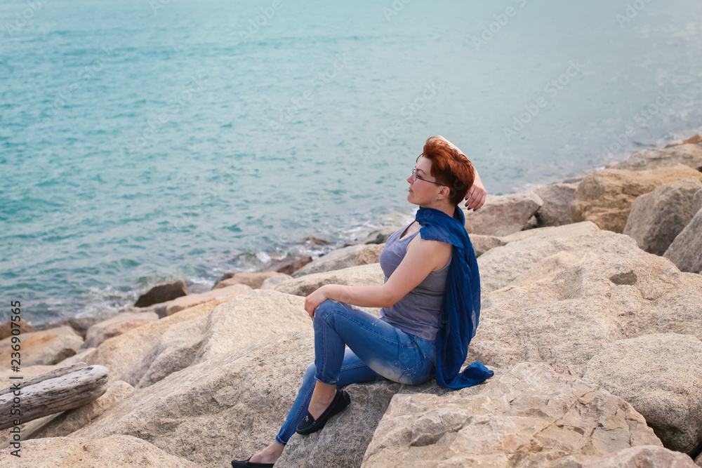 Adult caucasian women sit on rocky beach with blue neckchief relaxing and thinking about something