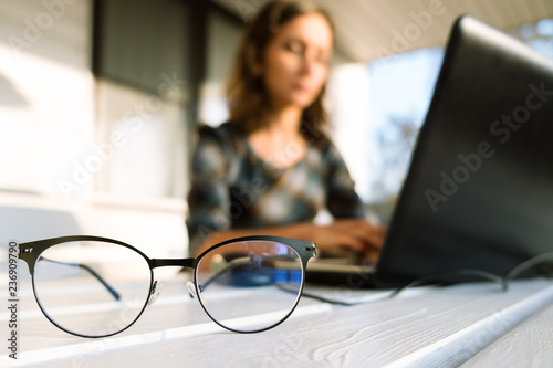 Young brunette woman sitting on outdoor terrace and using laptop. Hands on keyboard. Selective focus on eyeglasses. Home office, online shopping concept.